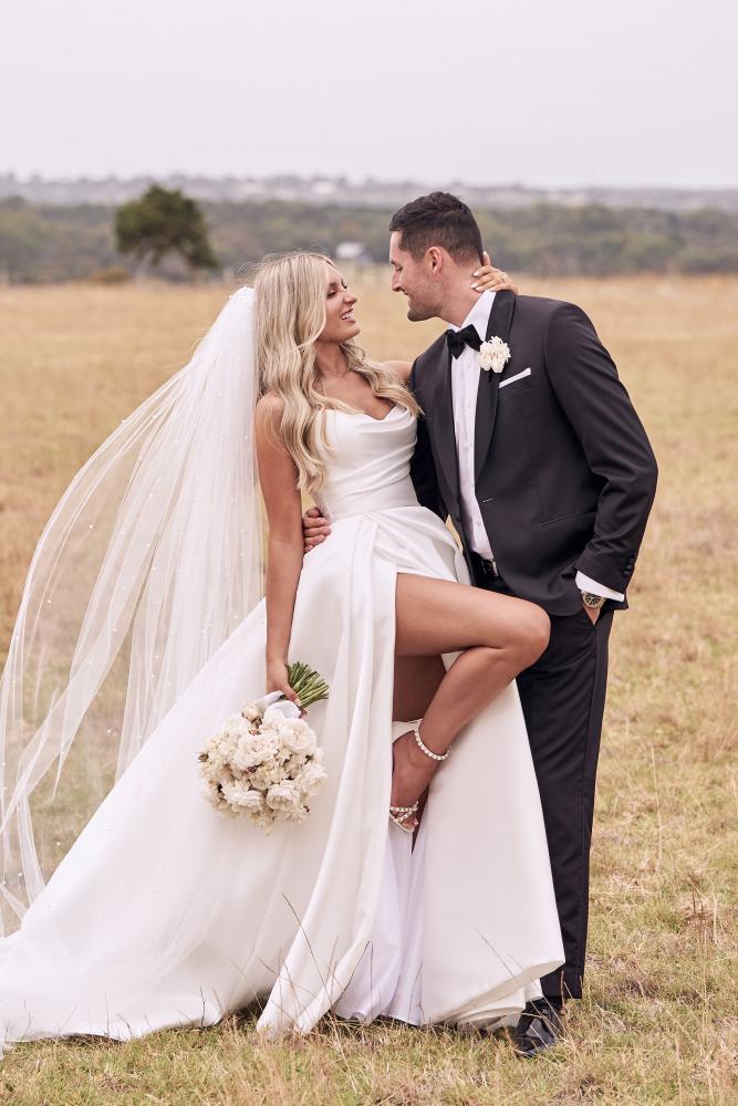 Bride wearing strapless Chantelle ballgown with sweetheart bodice and thigh slit, holding bouquet, with groom in black tuxedo outdoors