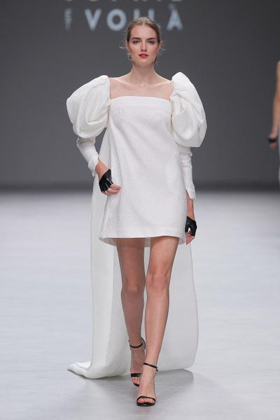 Model wearing the Dune white mini dress with puffed sleeves and a long train walking on a runway