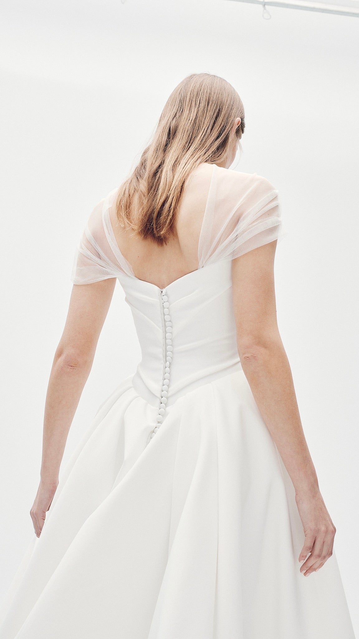 Model wearing the Aliet white strapless ballgown with extended corset and sheer draped sleeves, back view in minimal setting