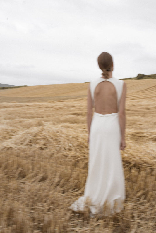 Model wearing the Bhirsty white bridal dress with a slim fit and open back standing in a wheat field
