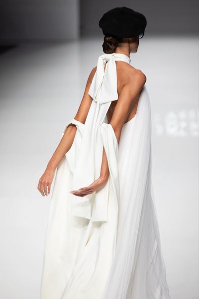 Model wearing the Campanille white flowing halter dress with large bow back detail and black beret on runway
