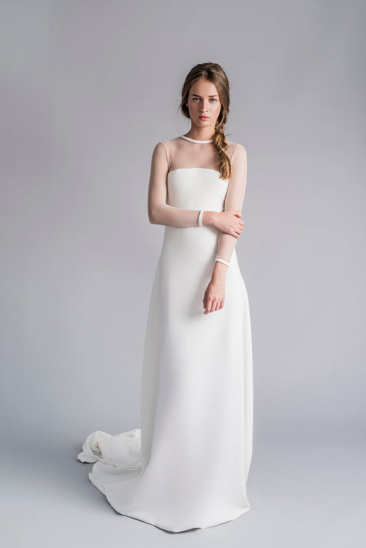 Model wearing the Candice white bridal gown with sheer long sleeves and a flowing train against a gray background
