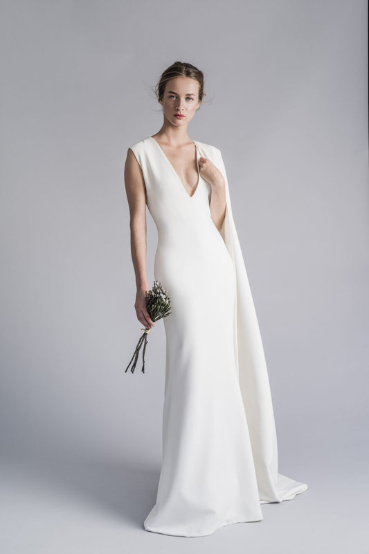 Model wearing the Catherina white gown with deep V neckline and cape detail holding a small bouquet on gray background