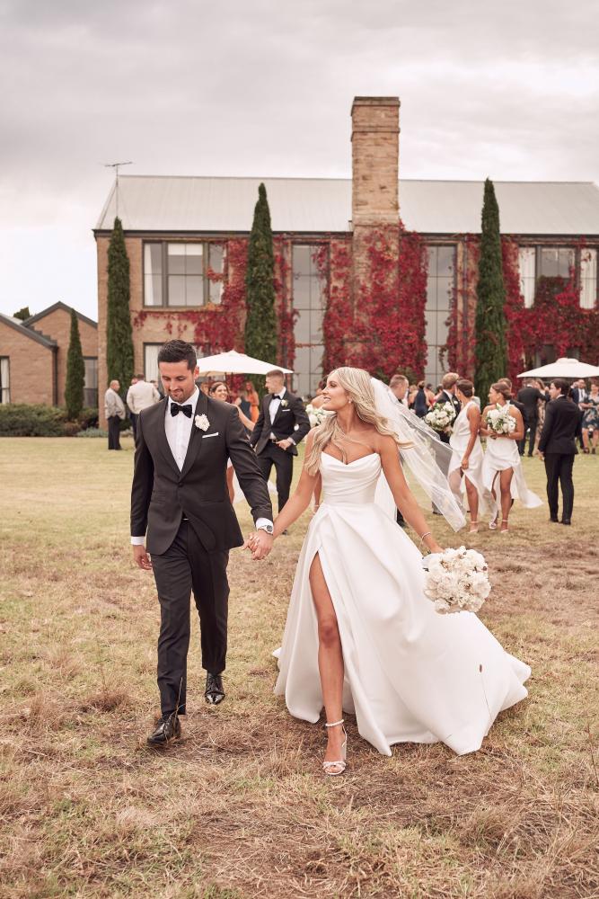 Bride wearing the Chantelle strapless ballgown with sweetheart bodice and slit skirt holding bouquet walking with groom outdoors