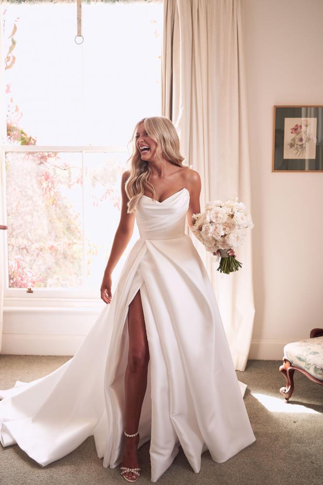 Woman in a white wedding dress with a high slit, holding a bouquet in a bright room.