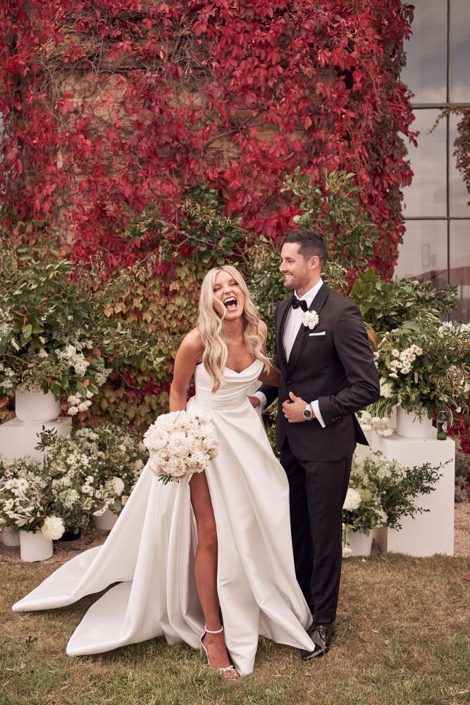 Man and woman in formal attire standing in front of a floral backdrop with red leaves.