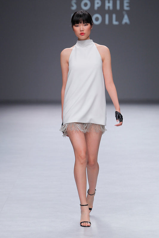 Model wearing the Daniella white halter dress with fringe hem and black glove walking on runway in Chicago setting