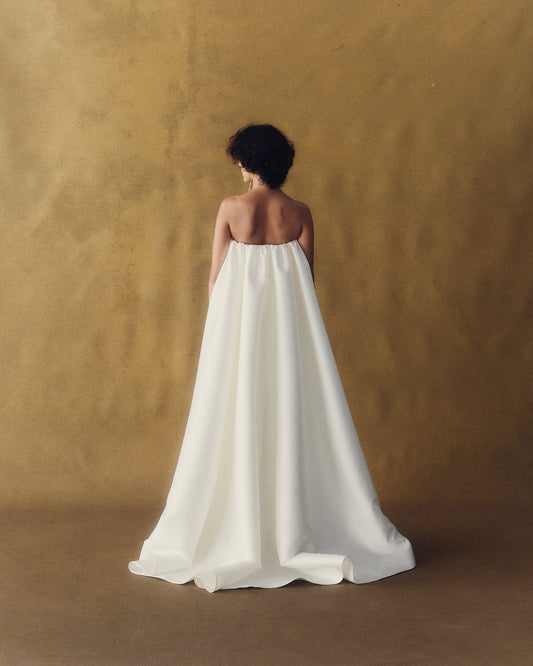 Model wearing the strapless Daphne Train in lustrous white twill fabric against a textured brown backdrop