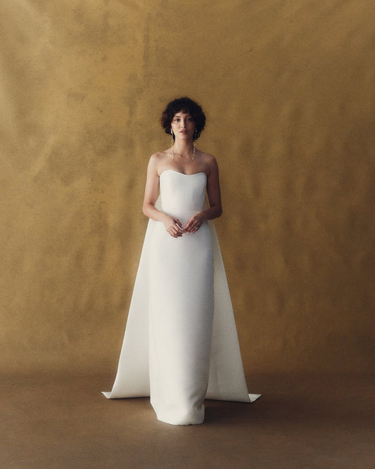 Model wearing the Eloise Tails white strapless gown with structured bodice and long trailing tails against a textured backdrop