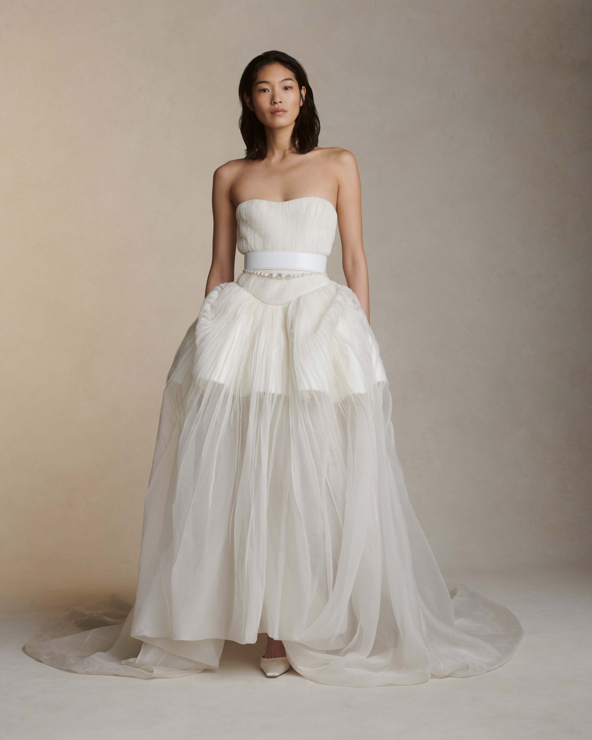 Woman wearing a strapless white wedding dress with a layered skirt against a neutral background