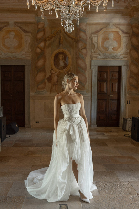 Model wearing Emory silk organza strapless ball gown with pintucked bodice and sheer high-low skirt in historic room