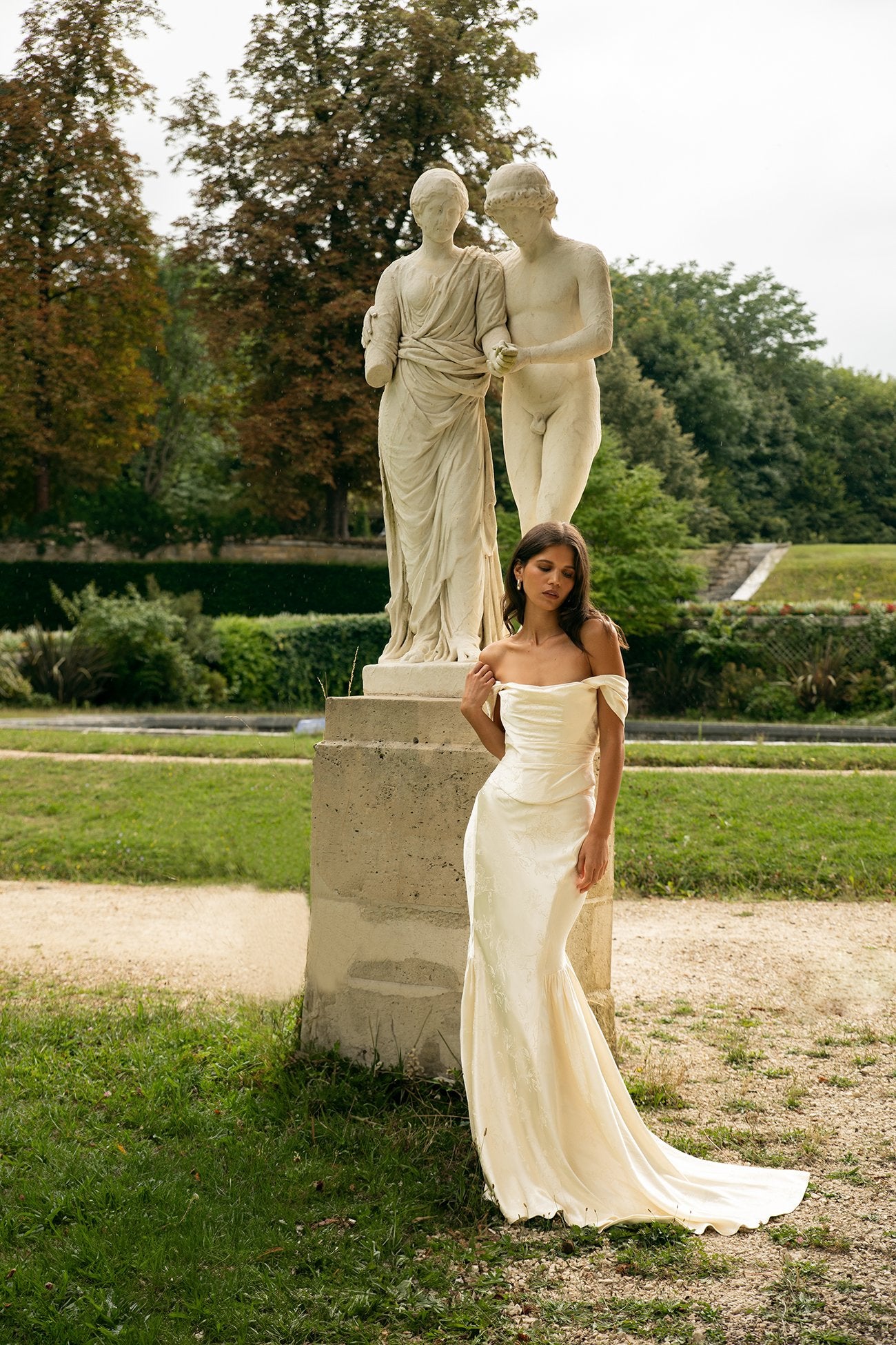 Woman in a white dress standing next to classical statues in a garden setting