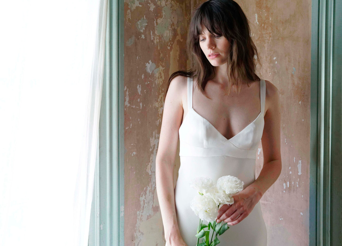 Woman in a white dress holding flowers against a textured wall.