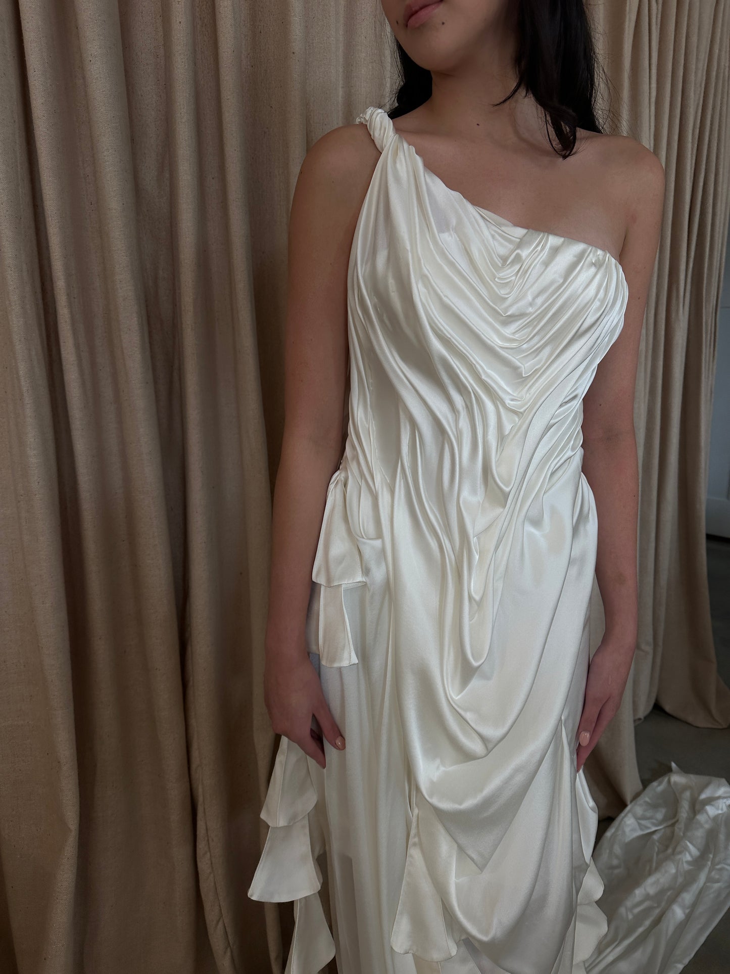 Model wearing the Silk Spring Awakening gown with intricate draping and one-shoulder design against a beige curtain backdrop