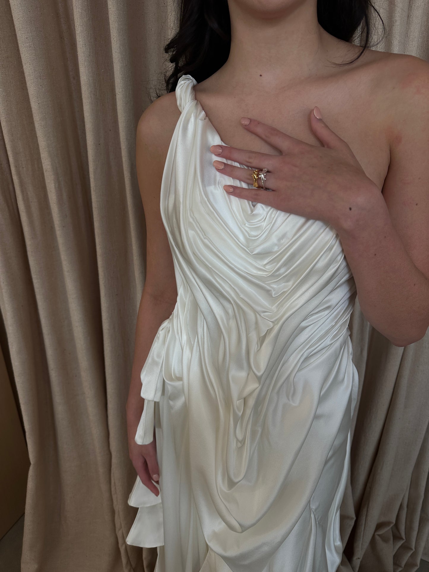 Model wearing the Silk Spring Awakening off-shoulder draped silk gown with fluid detailing against a beige curtain background