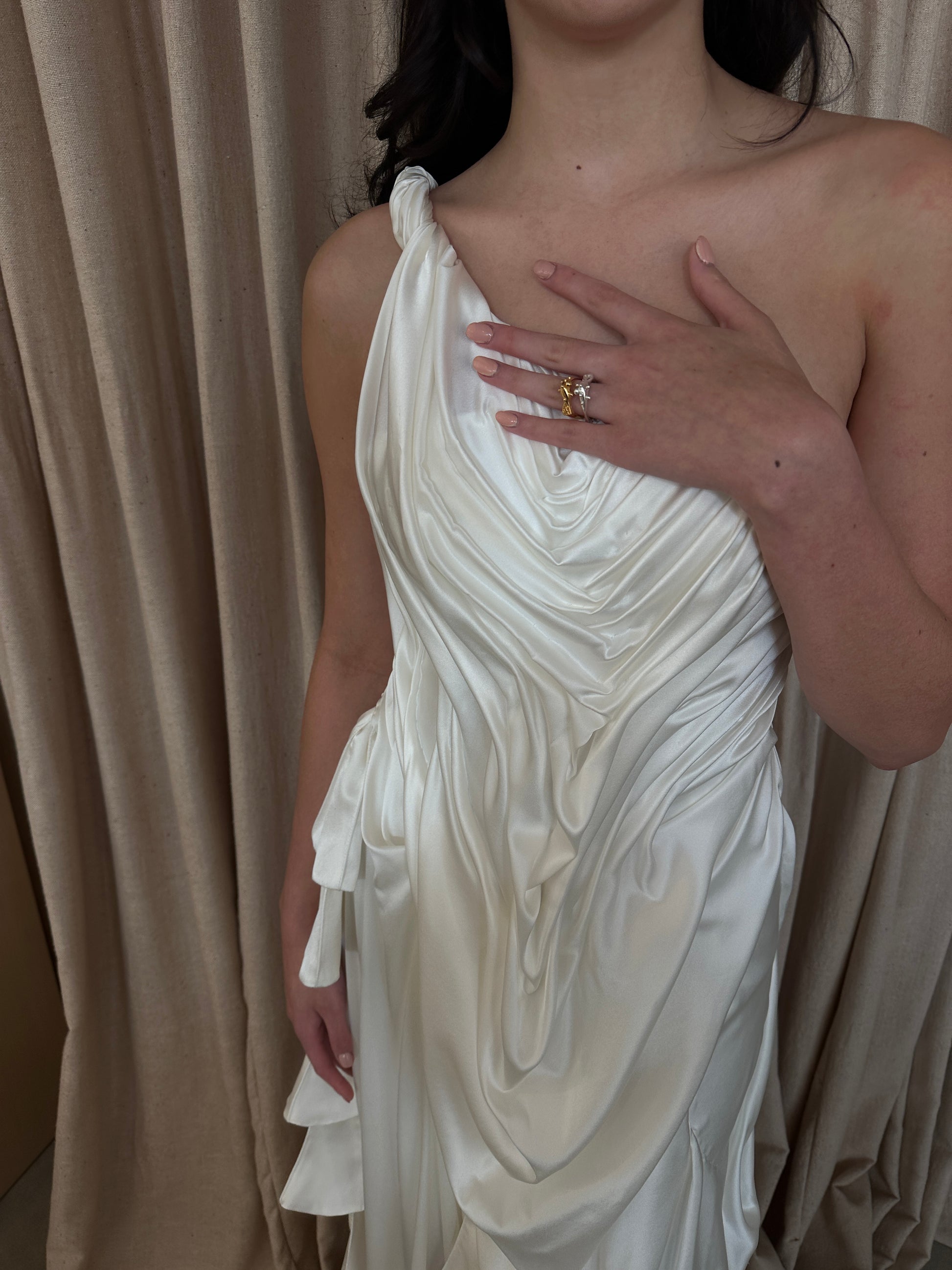 Model wearing the Silk Spring Awakening off-shoulder draped silk gown with fluid detailing against a beige curtain background