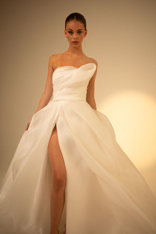 Model wearing a strapless white wedding dress with a high slit on a plain background