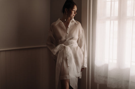 Woman wearing the Jones Mini sheer white button-up dress with a waist tie standing by a sunlit window