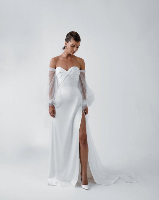 Model wearing the Karina white off-shoulder gown with rounded sweetheart neckline, corset waist, and thigh-high slit