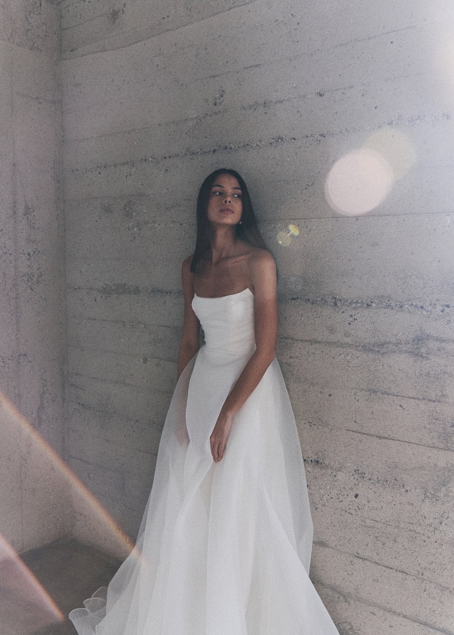 Model wearing the Liv white A-line dress with textured organza overlay standing against a concrete wall with sunlight flare