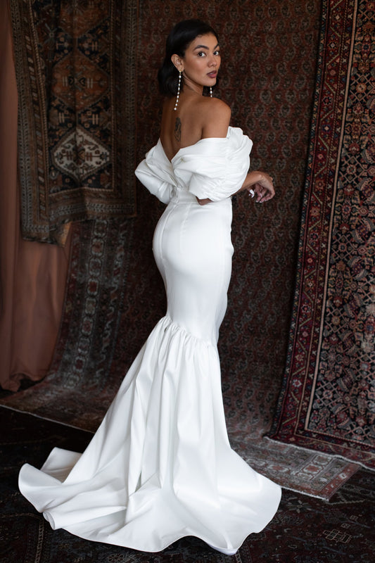 Model wearing the Larkin white off-shoulder skirt with puff sleeves and dramatic mermaid train against patterned rug backdrop