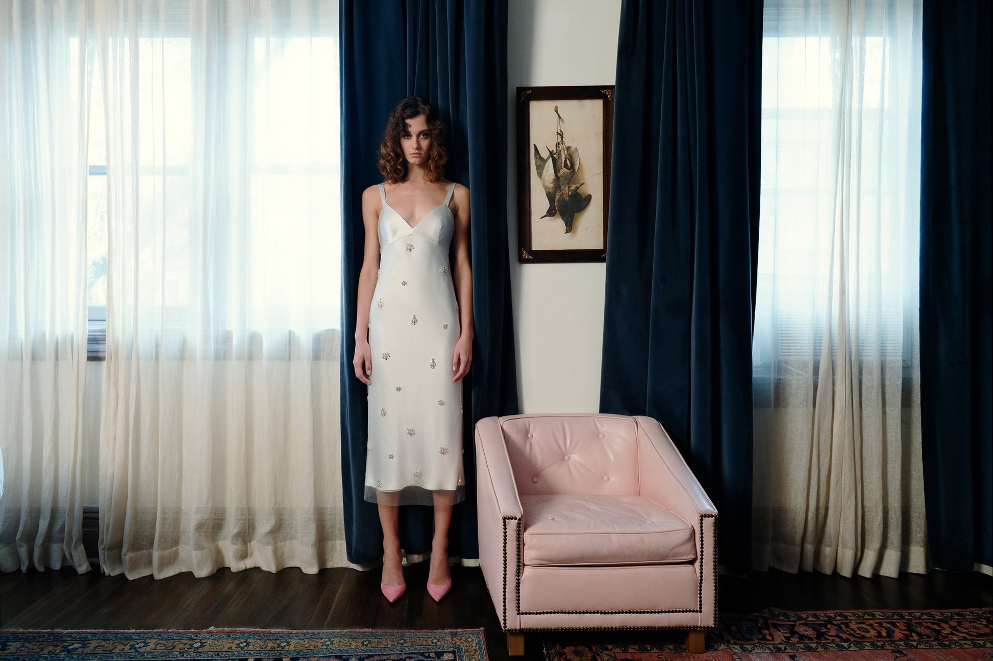 Model wearing the Lotus ¾ slip dress with jewel beaded tulle overlay in satin back crepe standing by navy curtains