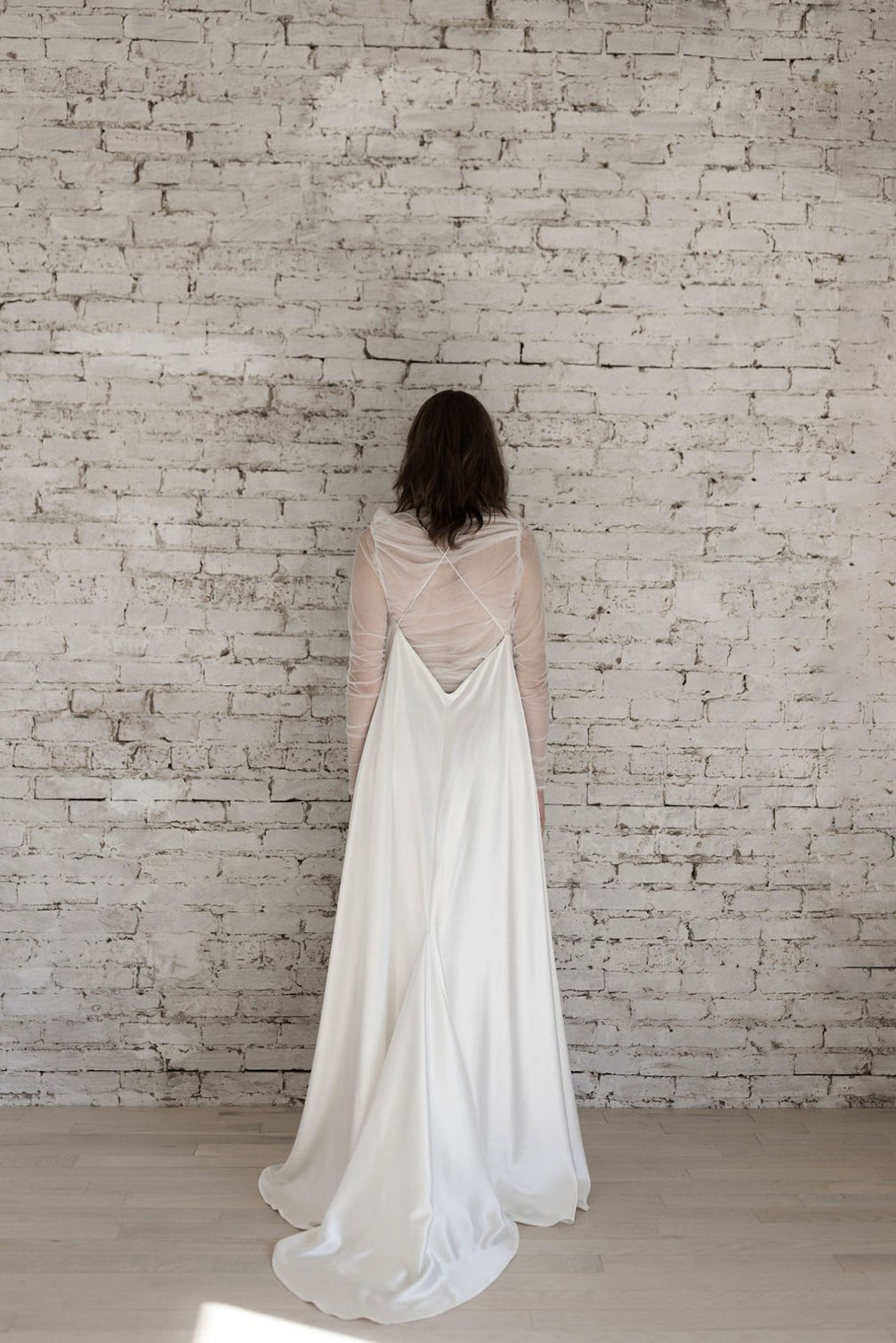 Woman in a flowing white dress standing against a light brick wall.