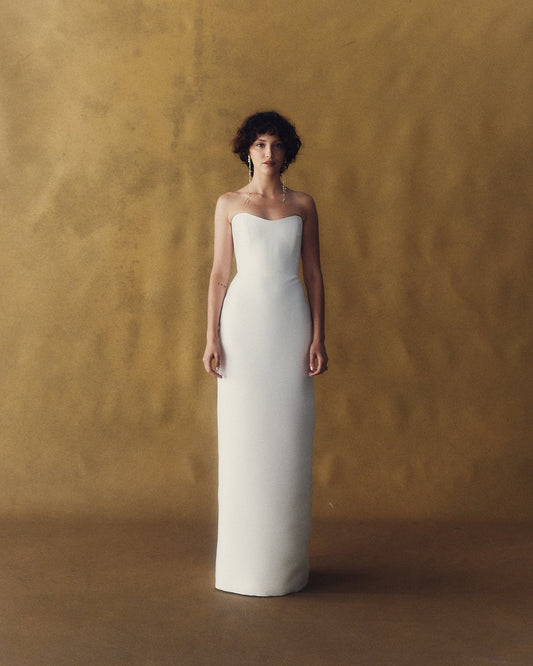 Model wearing the Mariska minimalist white strapless gown with a straight neckline against a golden textured backdrop