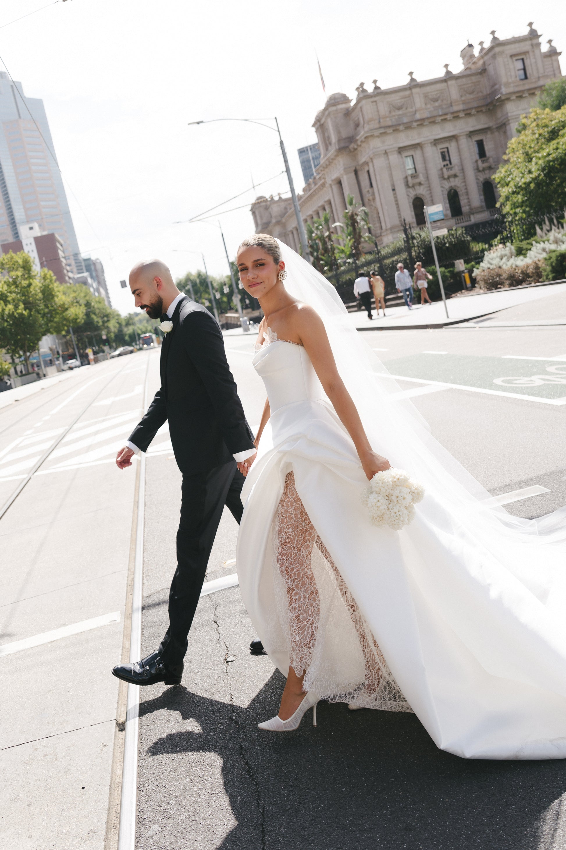 Man and woman in wedding attire walking on a city street with a large building in the background.