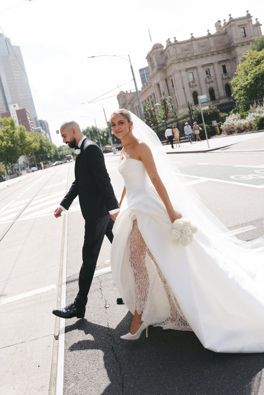 Man and woman in wedding attire walking on a city street with a large building in the background.