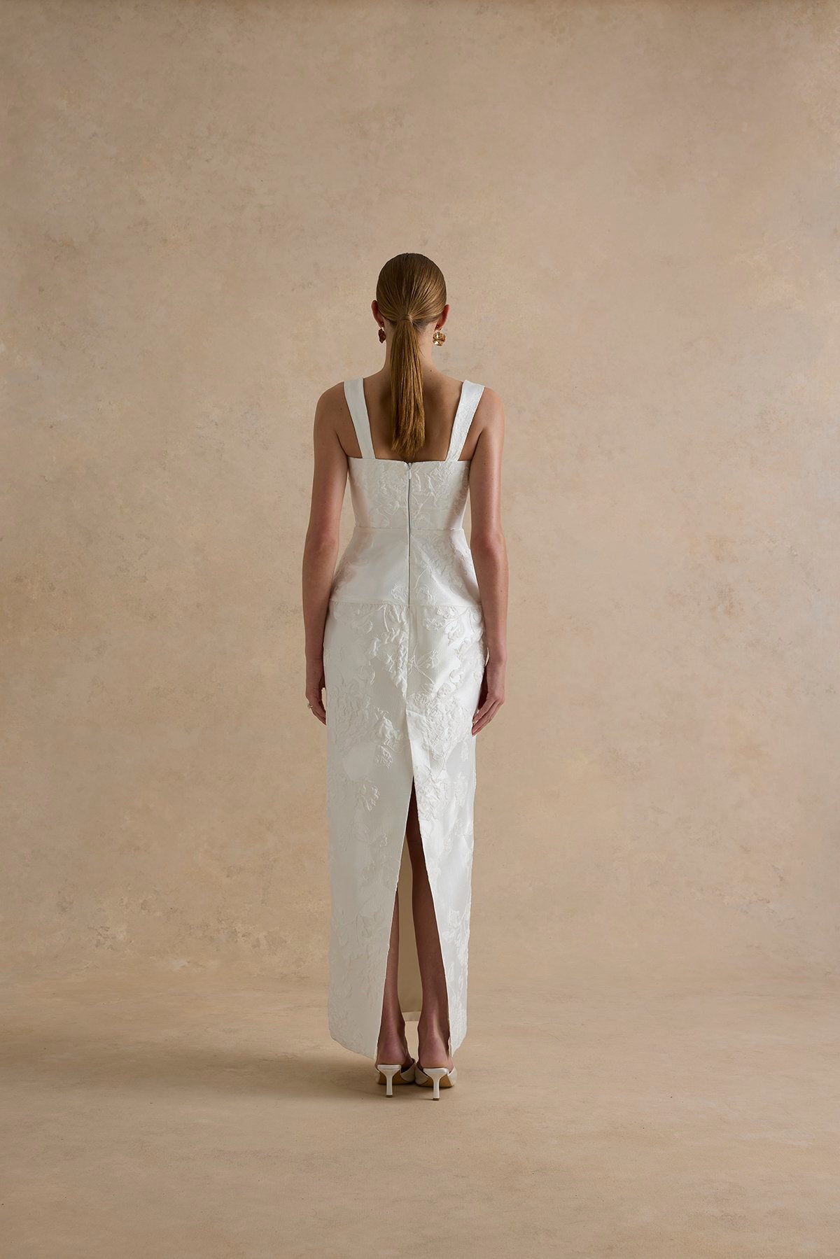 Woman wearing a white dress with a slit, standing against a beige wall.