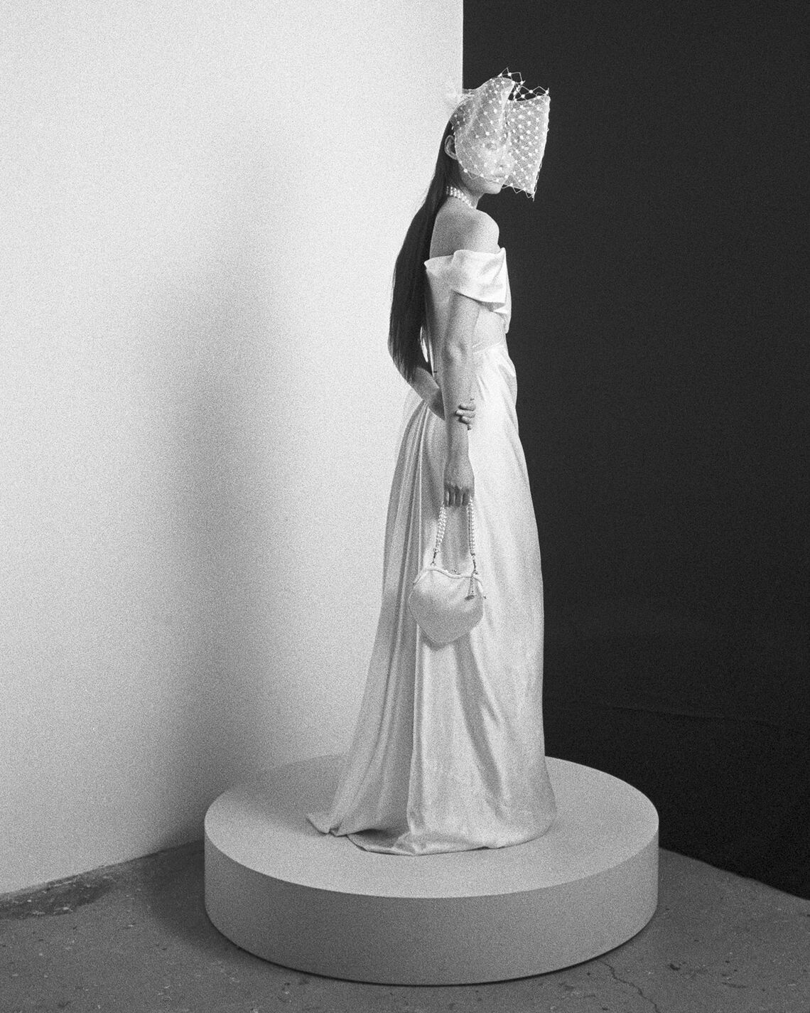 Model wearing the Nova Cocotte off-shoulder gown with corset waist and veiled headpiece on a round platform in black and white