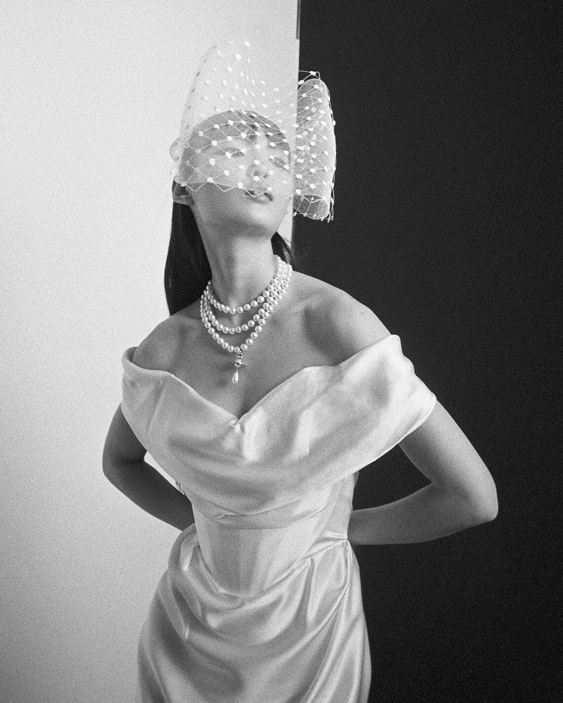 Black and white photo of model wearing the Nova Cocotte off-shoulder gown with pearl necklace and netted veil against split background