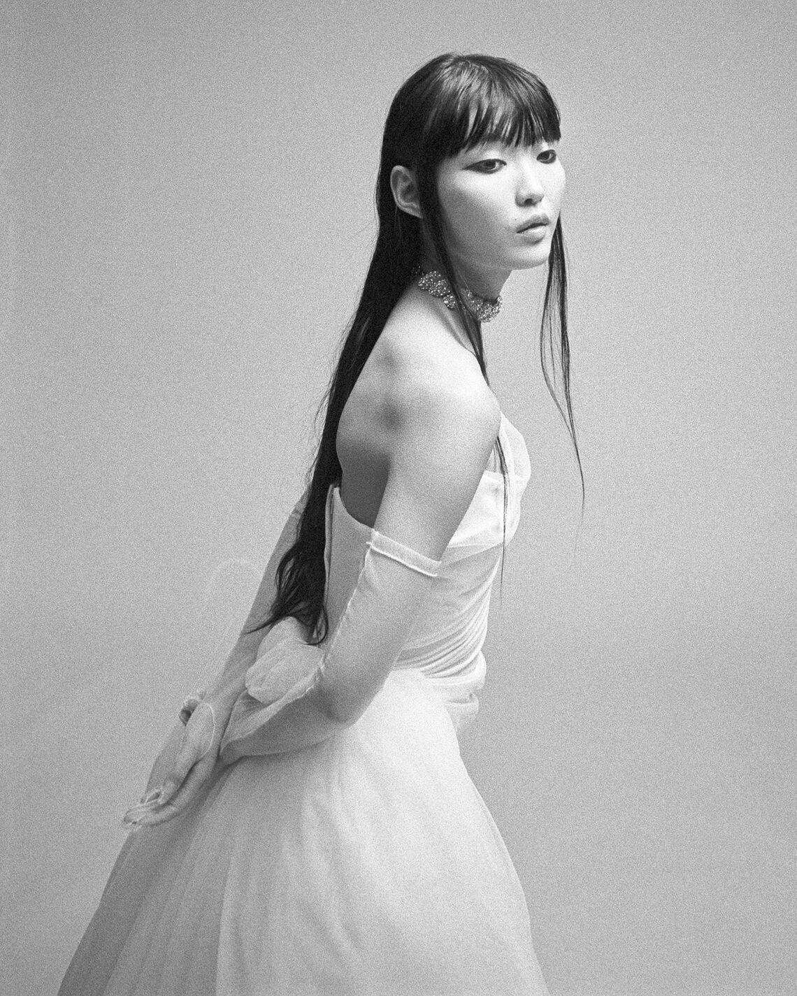 Black and white photo of model wearing the off-shoulder Nova Rhea gown with corset detailing and sheer gloves against a plain background