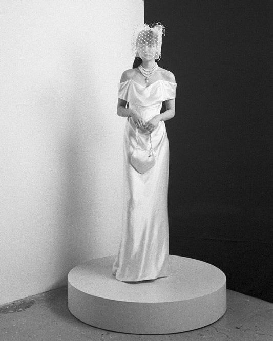 Black and white photo of model wearing Nova Cocotte off-shoulder gown with pearl necklace and dotted veil standing on round pedestal