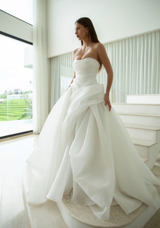 Woman in a strapless white wedding gown standing in a bright room with large windows.