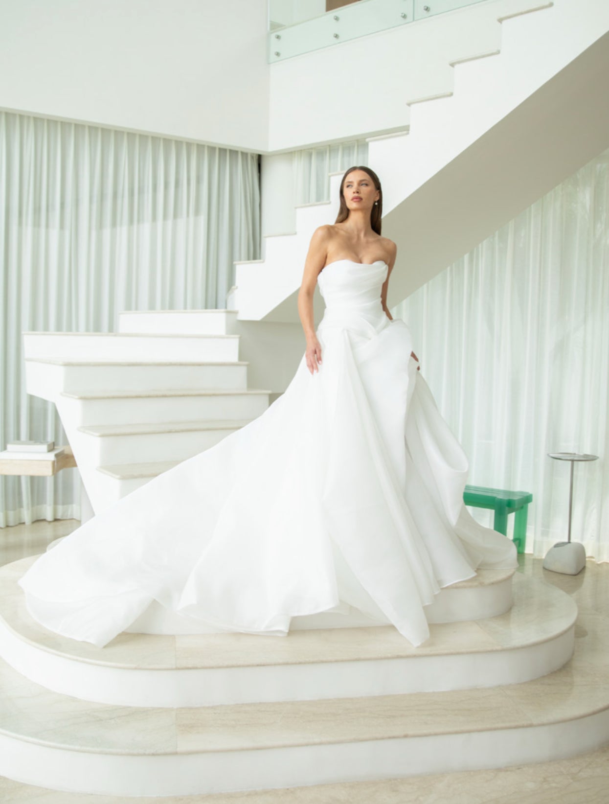 Woman in a white strapless gown standing on a staircase with white walls and green chair in the background.