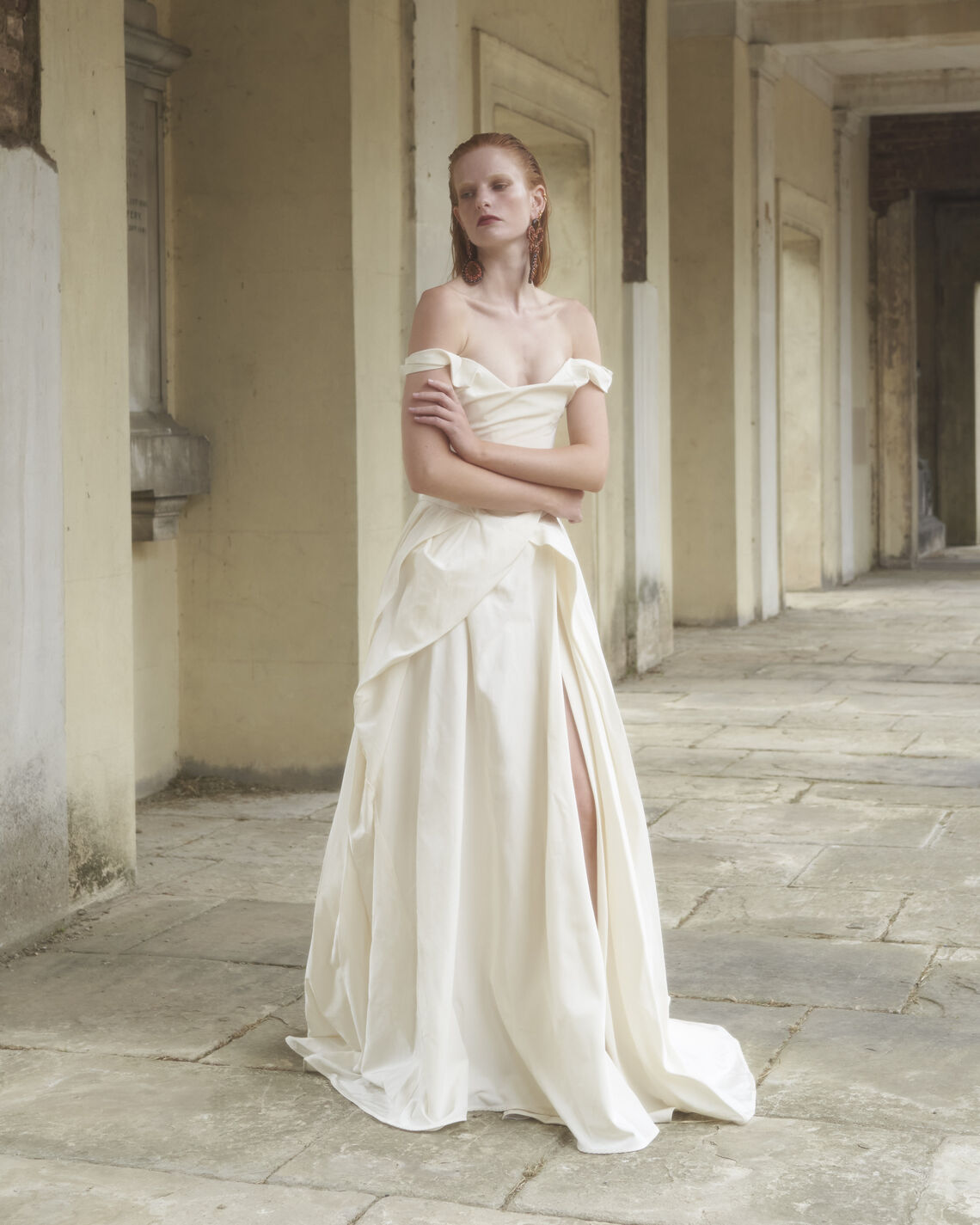 Model wearing the off-shoulder Tabitha gown with draped bodice and thigh slit standing in an architectural stone corridor