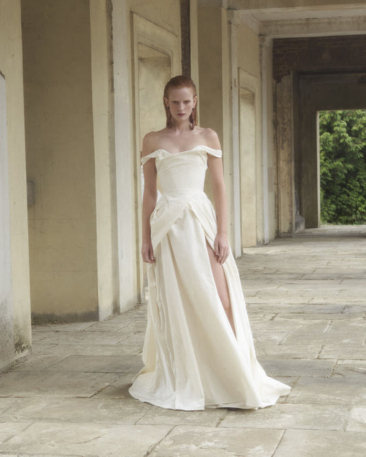 Model wearing the off-shoulder Tabitha white gown with draped skirt and thigh-high slit in a stone hallway setting