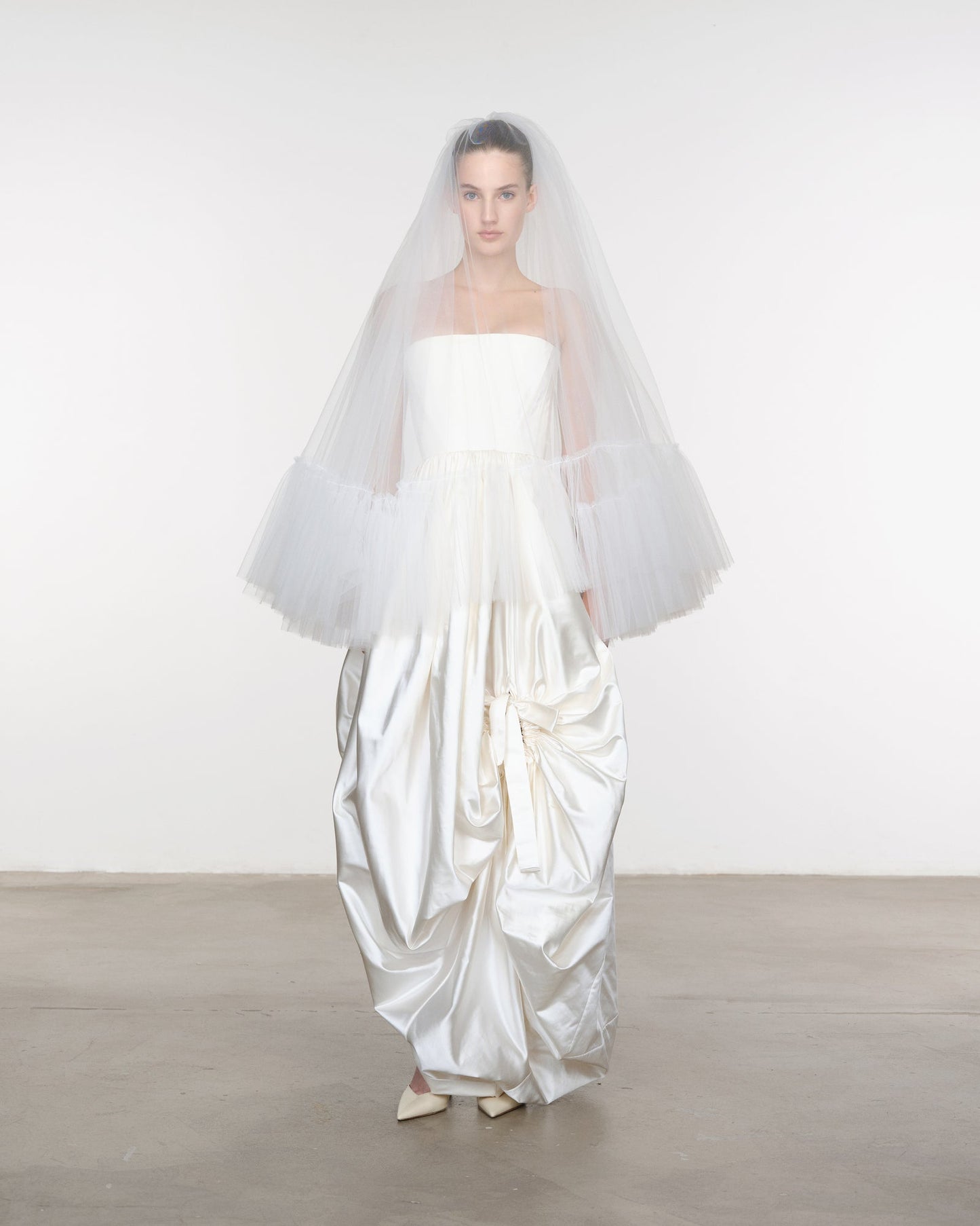 Model wearing a white wedding dress with a long train and veil against a plain background