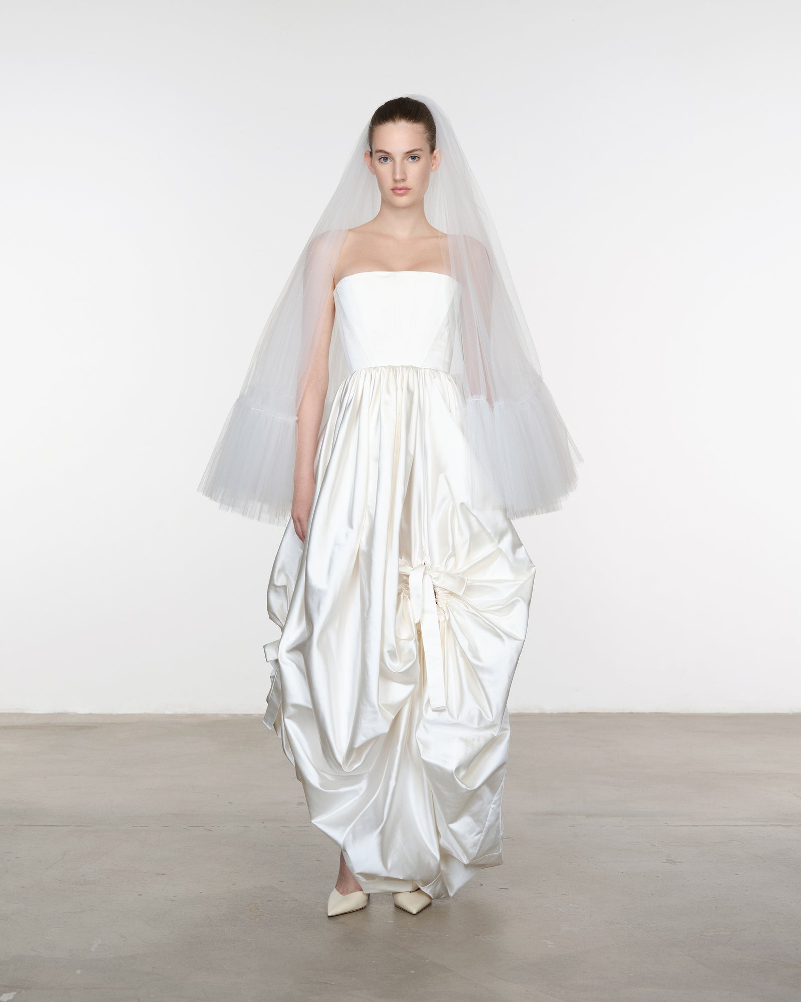 Model wearing a strapless white wedding dress with a flowing skirt and veil against a plain background