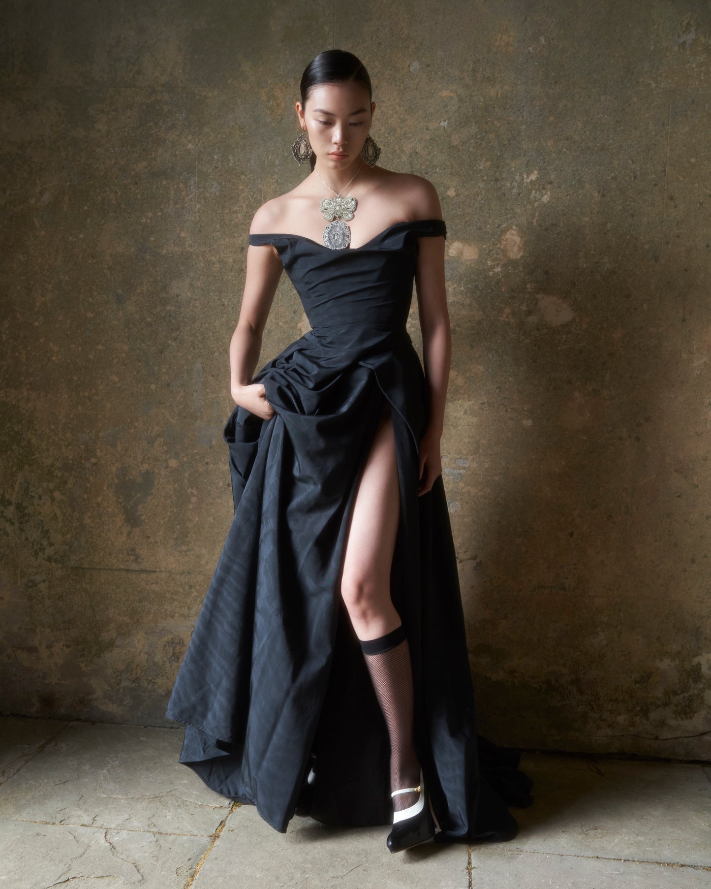 Model wearing the Tabitha black off-shoulder gown with thigh-high slit and sculpted draping against textured wall background