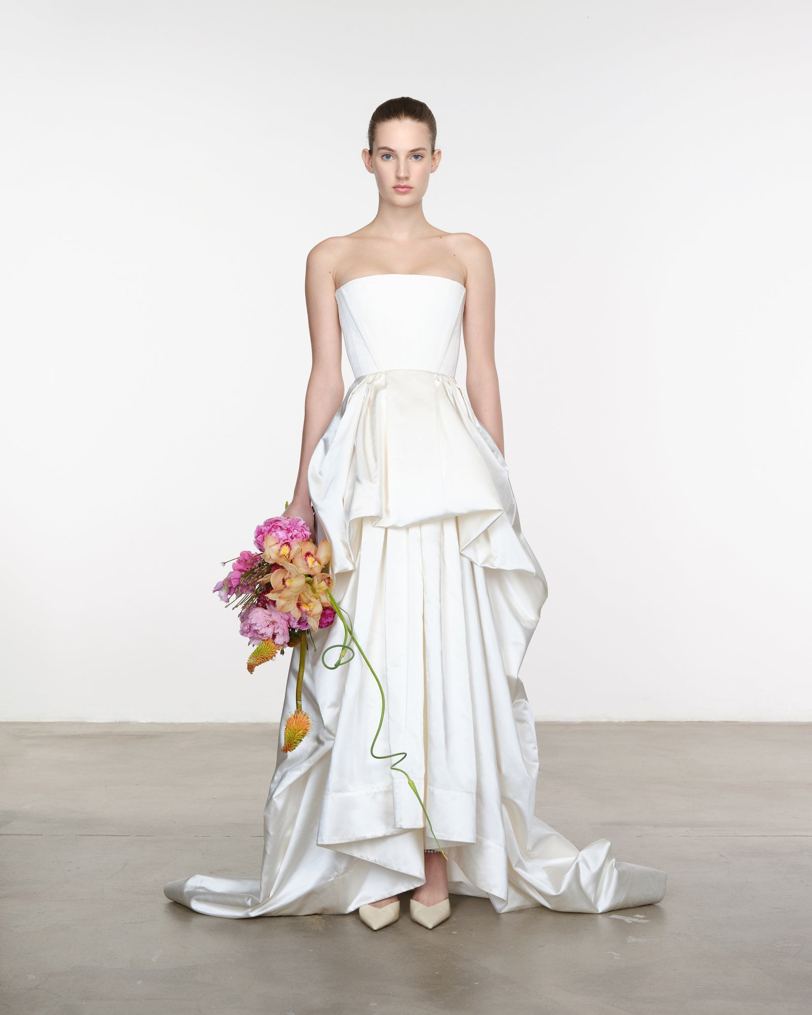Model wearing a white strapless gown with a ruffled skirt, holding a bouquet of flowers against a plain background.